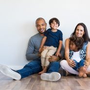 A couple sits with their two children against a white wall smiling